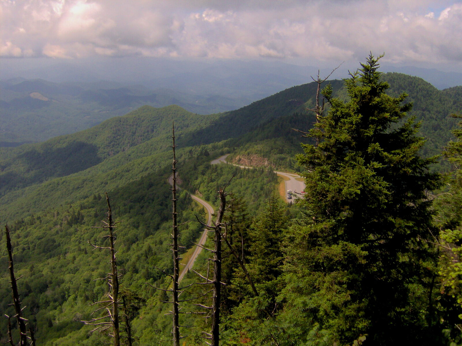 An overlook on the Blue Ridge Parkway, with the road curving away through forested mountains under a cloudy sky.