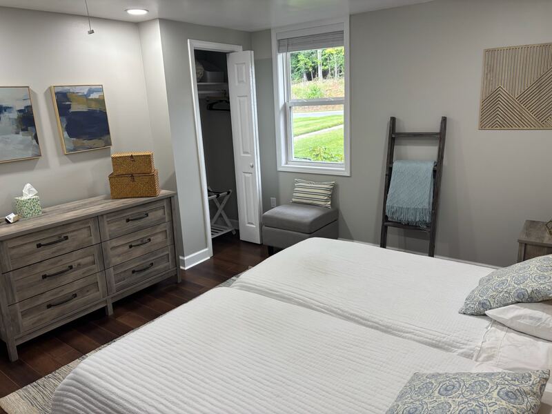 A bedroom with two queen beds in white bedding, a grey-washed dresser, a decorative ladder with a throw blanket, and a pair of abstract prints in blues.